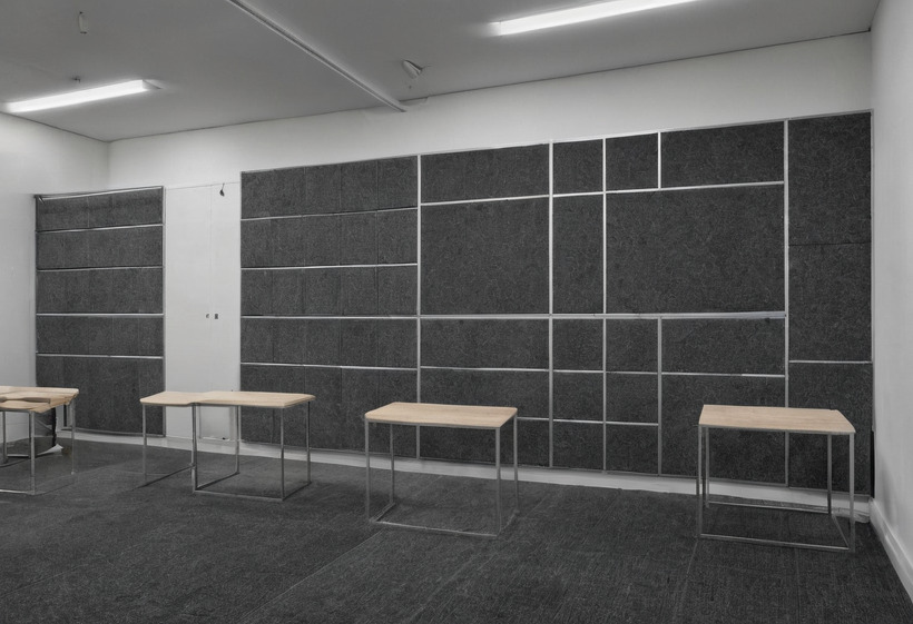 A wall of dark grey pin-up panels in an aluminium grid with four pale timber console tables standing empty against it on a grey carpet floor.