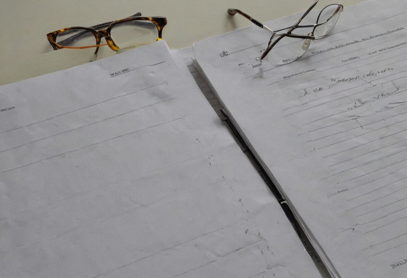 Two large ruled sheets of handwritten paper lying side by side on a desk, a slim dark pen between them and a pair of glasses resting at each top corner.